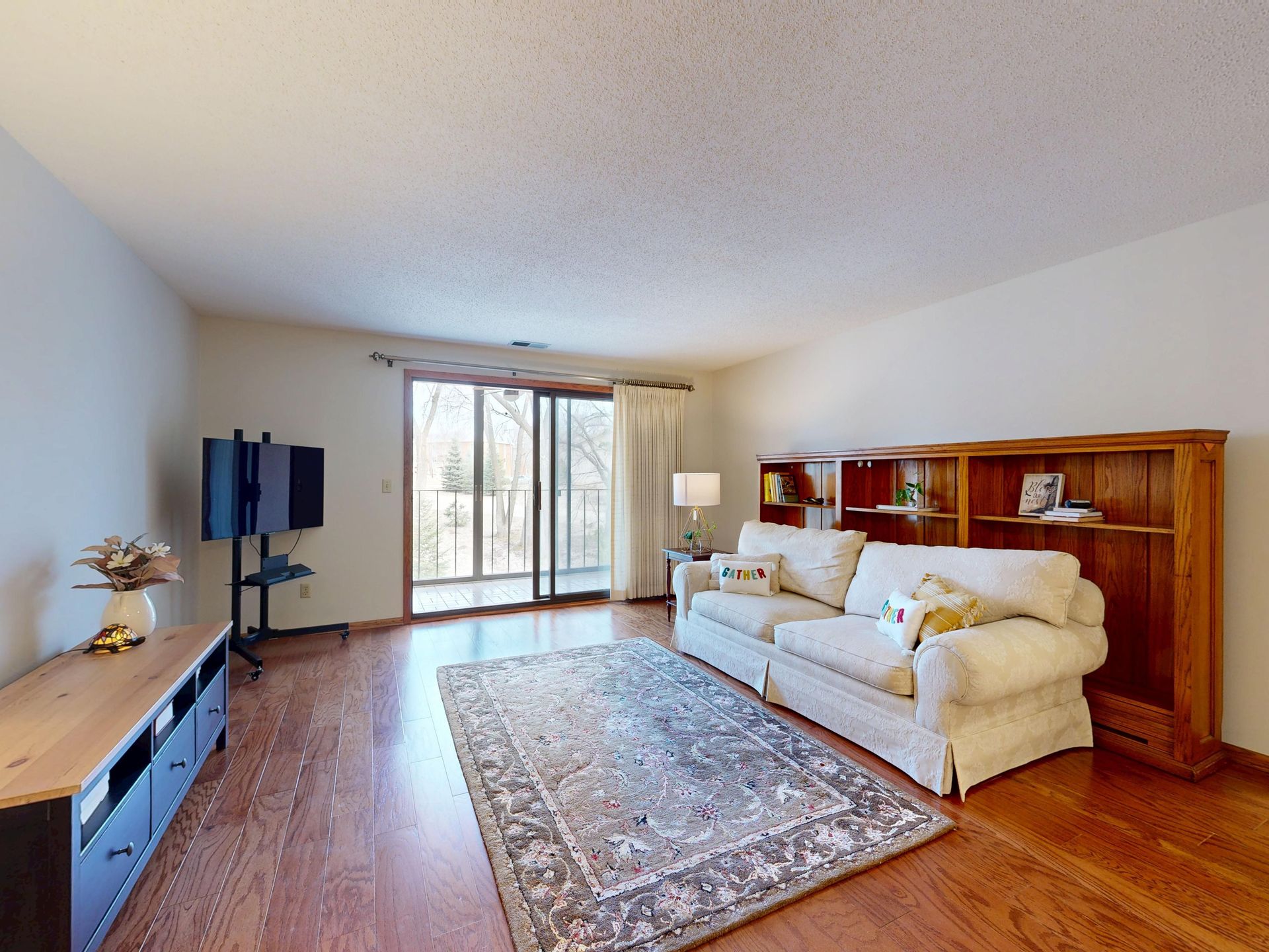 Spacious living room with sofa, TV, and balcony view, featuring hardwood flooring and wooden furniture.